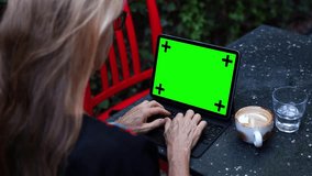 Over shoulder view of digital nomad woman wearing ethnic clothes in tropical setting having video chat online with green screen laptop computer.