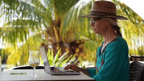 Attractive happy mature elderly woman digital nomad with straw hat having business video chat on a laptop computer while sitting at a table in tropics with palm trees behind her.