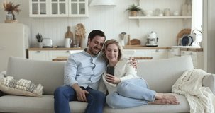 Happy millennial married couple relaxing on couch, talking on video call on mobile phone, using digital gadget for communication, hugging, speaking, smiling, laughing