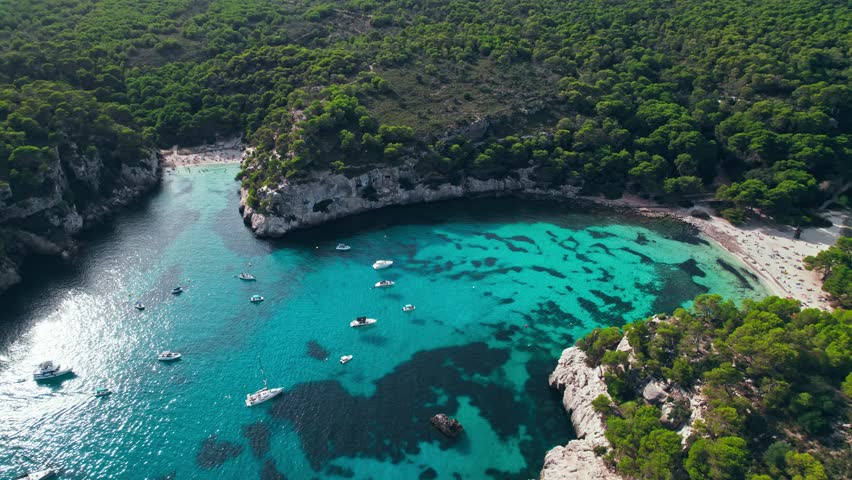 Aerial drone view of people having a good time by the beach with turquoise water. Tourists enjoy Cala Macarelleta and Macarella on the Balearic Islands, Spain, swimming and sailing yachts and boats.