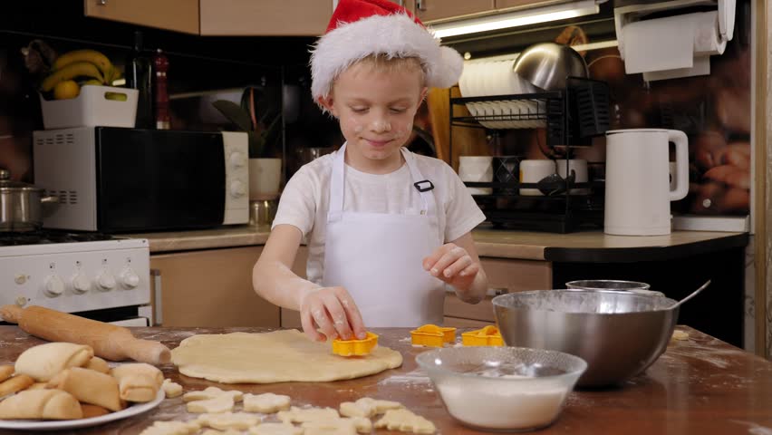 A cute boy in a Christmas hat cuts out various cookies using cookie cutters at home in the kitchen. 