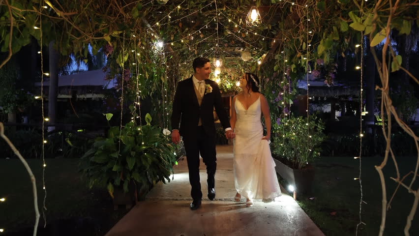 Stunning bride and groom heading to the wedding party in cinematic venue. Newlyweds walking along beautifully decorated garden with yellow lights. High quality FullHD footage Royalty-Free Stock Footage #1101496565