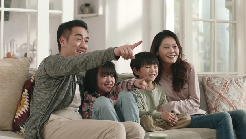 asian family with two children sitting on couch at home watching tv