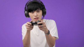 Asian young man in white t-shirt and playing video games using joysticks with headphones on voilet background isolated.