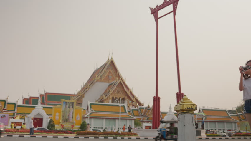 Young female traveler taking a photo of the Giant Swing and Wat Suthat temple in Bangkok, Thailand. Solo sightseeing and urban heritage exploration.