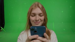 Wide-angle video of a white race cheerful red-haired girl looking at something on a smartphone and smiling on green screen background