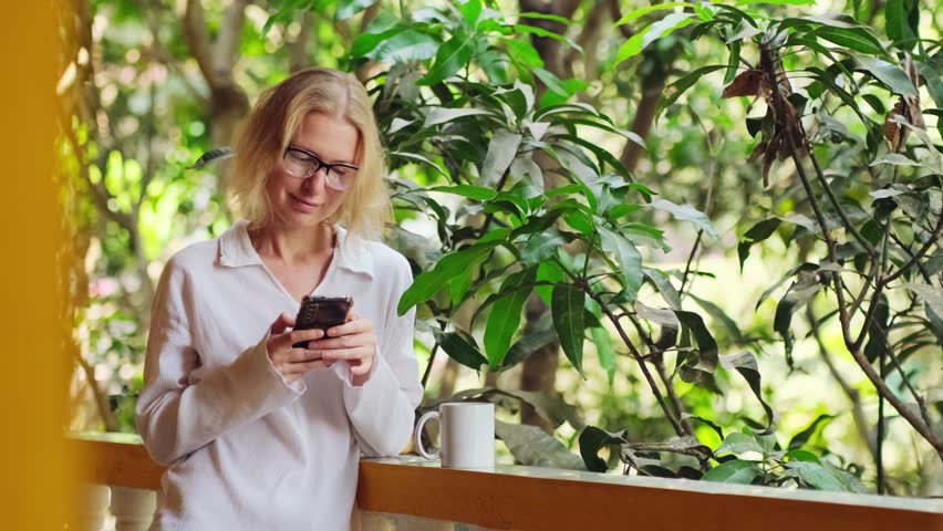 Caucasian woman is using her smartphone, drinking morning coffee on the balcony. Female blonde texting at the terrace outdoor. Good start to the day.