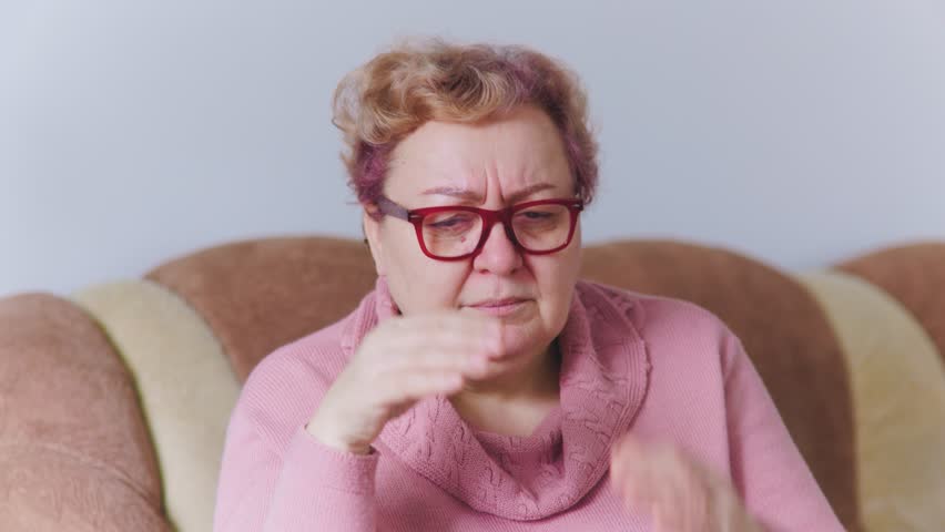 Elderly woman experiencing an intense headache, with her hands tightly clasped on her head, in her comfortable home | Shutterstock HD Video #1101521899