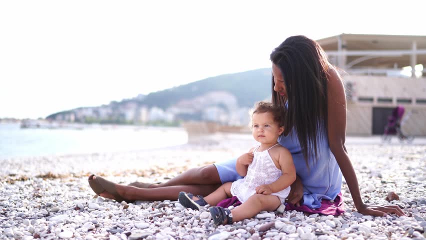 Mom plays pebbles with a little girl, sitting with her on a blanket on the beach