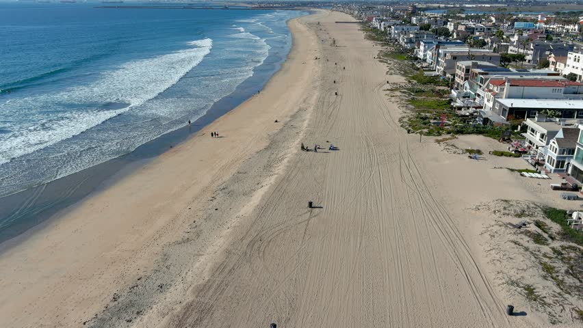 aerial footage flying over the coastline with blue ocean water, palm trees, grass and people walking along the sand beach at Sunset Beach in Huntington Beach California USA