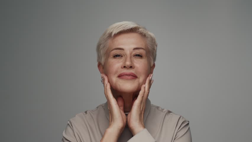 Portrait of adult woman looking at camera in colorful studio shot. Old female isolated alone on white background closeup. Headshot of aged person touches skin of healthy face by hands and smiles happy | Shutterstock HD Video #1101530425