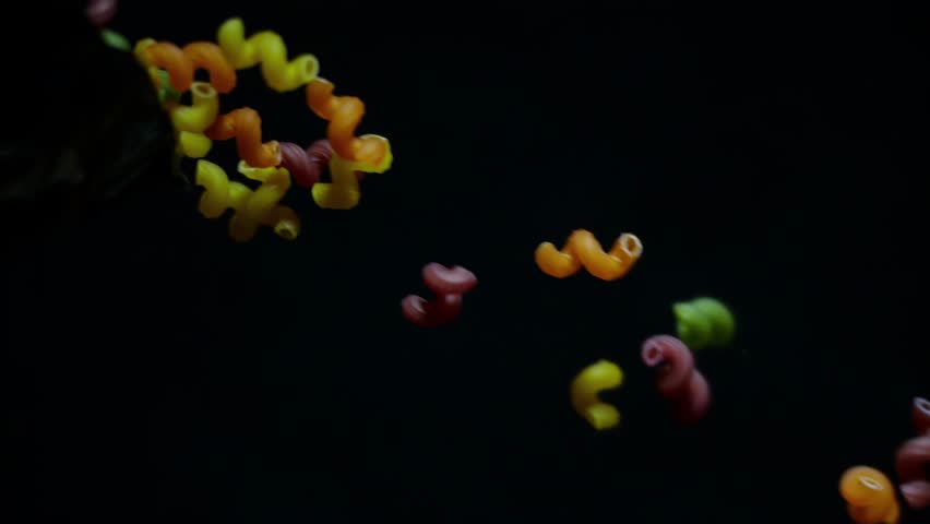 A lot of raw multi-colored pasta fall diagonally from a bag on a black background in slow motion
