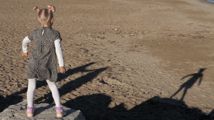 Blonde girl dances on the beach looking on her dancing shadow on the sand. Wide shot. Slow motion