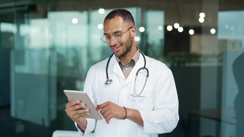 Portrait of a young smiling doctor using a tablet while standing in a clinic. Handsome mixed race therapist in glasses and white coat looks at news on the Internet, writes back messages, reads email Royalty-Free Stock Footage #1101539795