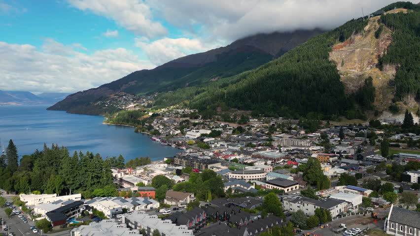 A lovely view from above the Queenstown city centre. Beautiful landscape includes a harbour with sky blue water, mountains with tops hidden in the clouds, and a city lying on the coast. Royalty-Free Stock Footage #1101541635