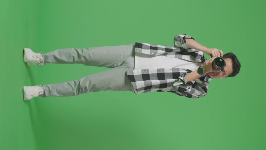 Full Body Of Asian Photographer Using A Camera Taking Pictures And Smiling Touching His Chest While Standing On Green Screen Background In The Studio
