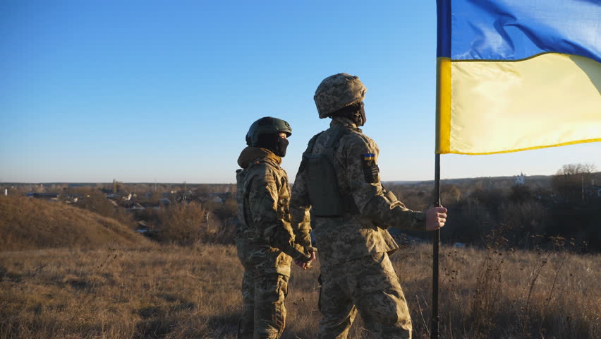 Young soldiers in camouflage uniform stands with a waving Ukraine flag at meadow against sunset. Military couple of ukrainian army lifted banner holding hands of each other at field. Dolly shot Royalty-Free Stock Footage #1101546997