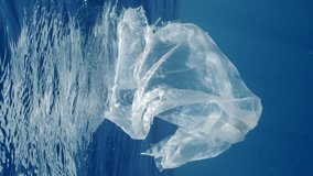Vertical video, Disposable acrylic bag floating under waves in blue water, Slow motion. Old torn plastic bag drifts underwater on sunny day in sunbeams. Plastic pollution og Ocean