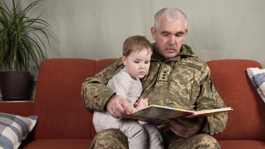 A man in Ukrainian military uniform reads a book to a little boy on the couch at home. Military grandfather reads a book to his grandson. Translation: Armed Forces of Ukraine