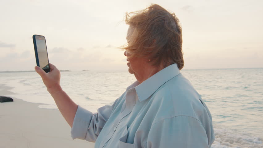 Elderly woman films video on her smartphone on the tropical beach at sunset | Shutterstock HD Video #1101552687