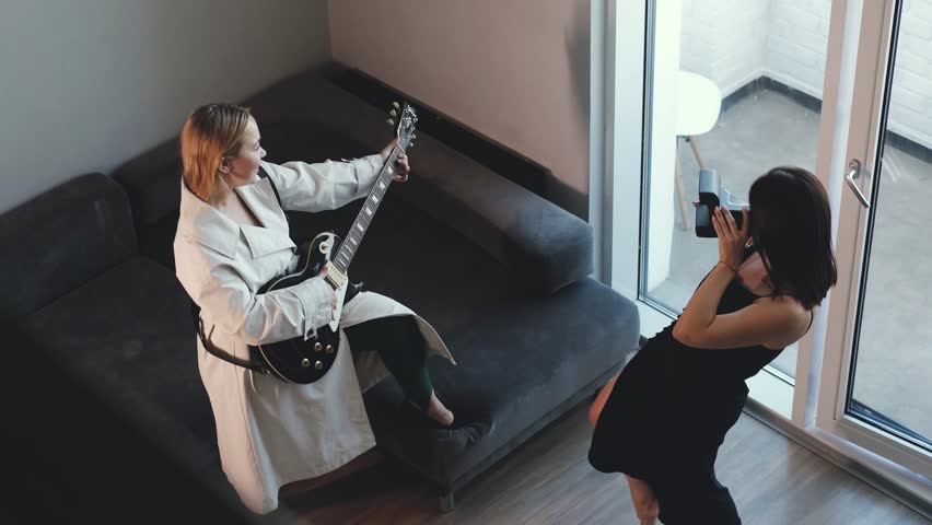 Two best friends have fun together. Young beautiful woman plays the guitar and sings a song at home. Her friend pretends to take a picture of her with a serious face. Women play a successful life and