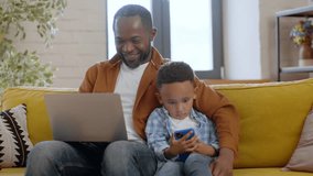 Gadgets addiction problem in modern families. Tracking shot portrait of young african american father and his little son using laptop and smartphone, sitting together at home, slow motion, free space