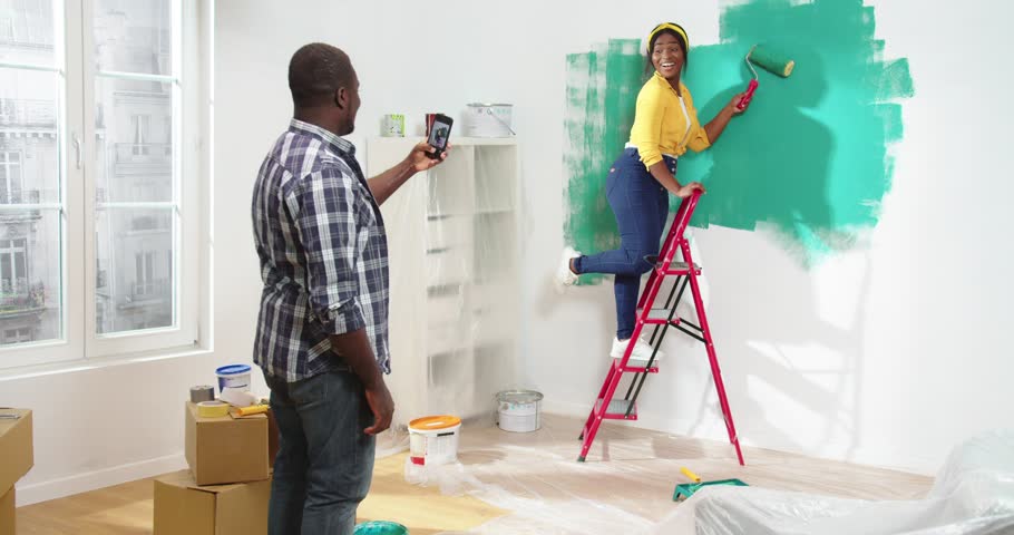 Loving handsome African-American man standing in flat during renovation taking picture of beautiful African woman painting walls with green paint posing holding roller standing on ladder. Royalty-Free Stock Footage #1101593927