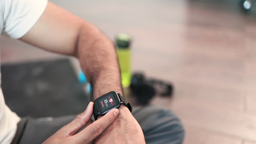 Close-up of young sporty indian man sitting on wooden floor and checking pulse on modern smartwatch with sports app after workout at home. Athletic male leading healthy lifestyle by training indoors.