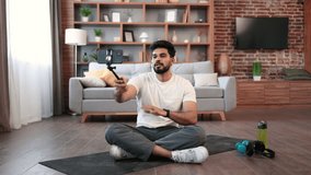 Active arabic male in cross-legged sitting pose having personal training online via cell phone on tripod in spacious room. Confident fitness blogger making live stream of morning workout using camera.