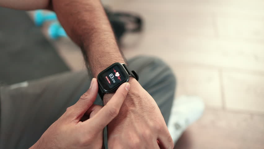 Close-up of young sporty indian man sitting on wooden floor and checking pulse on modern smartwatch with sports app after workout at home. Athletic male leading healthy lifestyle by training indoors. Royalty-Free Stock Footage #1101603139