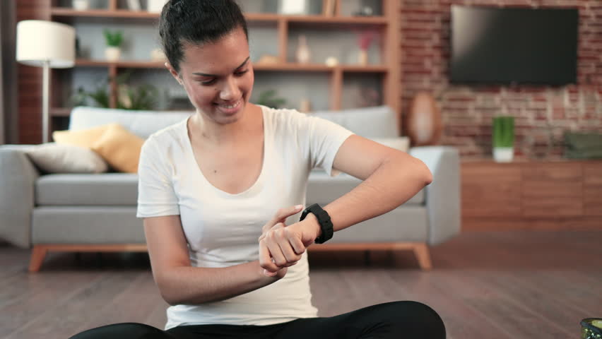 Young multiracial female looking at activity fitness tracker while relaxing on floor in apartment. Cheerful fit woman checking heart rate on modern smartwatch while having break from home workout.