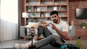 Young indian fitness expert sitting in front of phone camera on yoga mat near sports equipment for home workout. Active male blogger in gym wear recording home exercise for his online blog followers.