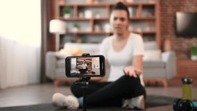 Selective focus of modern gadget on tripod recording active african american woman in sportswear talking at camera while resting on floor indoors. Personal coach guiding fitness training online.