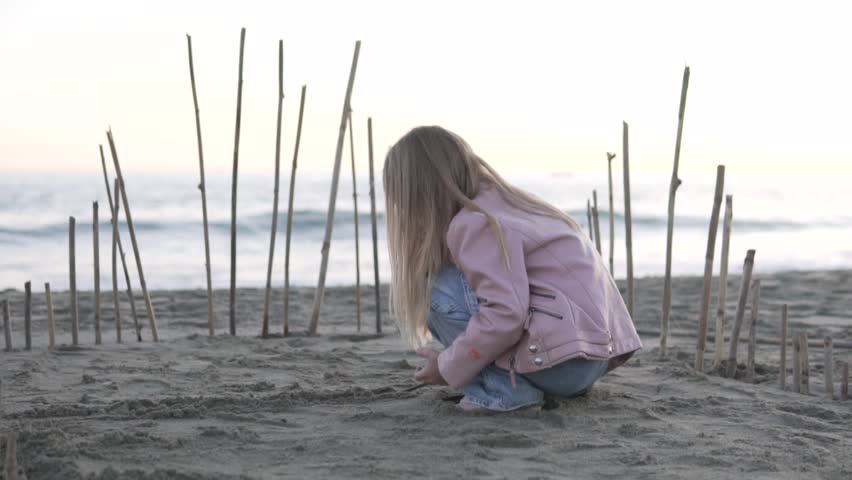A little girl in a trigger and jeans plays with sticks in the evening on the beach. A girl builds a hut on the beach, Turkey, Alanya | Shutterstock HD Video #1101606605
