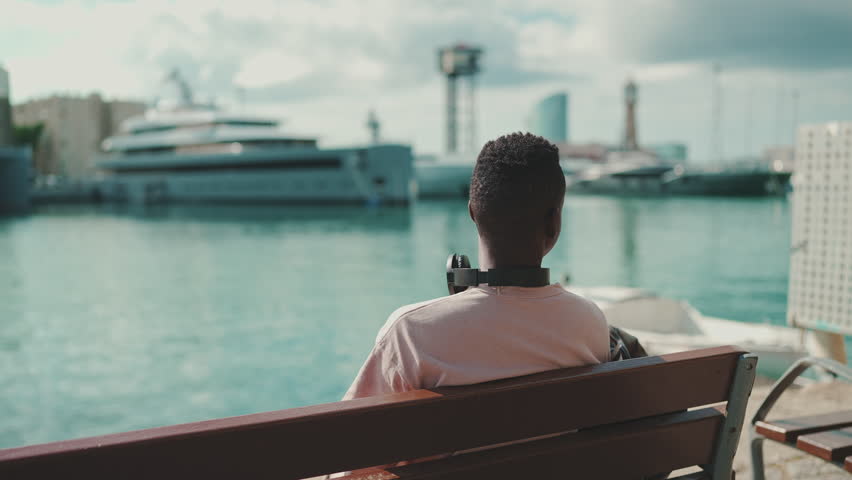 Young guy relaxes sitting on bench in the port on yachts and ships background. Back view