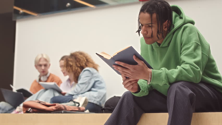 Slowmo of Biracial guy in oversized green hoodie reading book while waiting for class studying at college Royalty-Free Stock Footage #1101608545