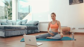 A young pregnant woman in sportswear sitting on a rubber mat in the living room at home doing yoga sports on video lessons on a laptop. Active lifestyle during pregnancy.