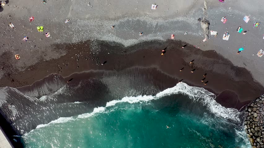 Straight down aerial drone footage of the beautiful town of Costa Adeje, Santa Cruz de Tenerife in Spain showing the Playa de Ajabo beach with people relaxing and having fun on the black sandy beach.