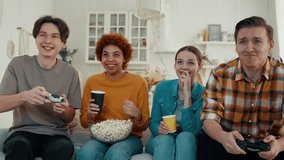 Company of multiethnic friends play video games at home. Two guys competing and two girls cheering them drinking and eating. Diverse people having fun hanging together. Leisure, entertainment concept.