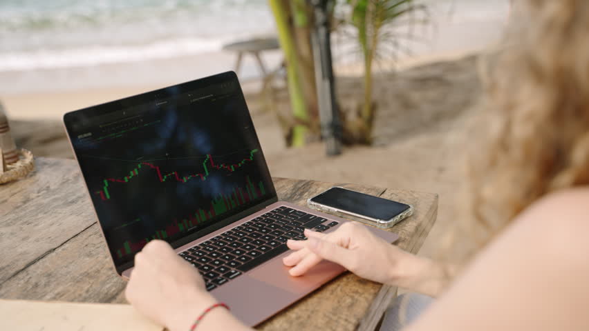 Female crypto trader at laptop checking candlestick charts online working remotely at outdoor tropical seaside cafe. Woman broker analyses cryptocurrency exchange rates on crypto stock by ocean | Shutterstock HD Video #1101613927