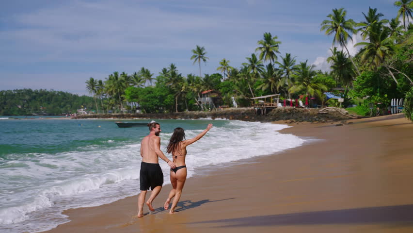 Young cheerful biracial couple in swimsuits holding hands running along ocean tide in waves splashing them slow motion shot. Multiracial happy man and woman run joyfully along sea in slowmo back view | Shutterstock HD Video #1101613939