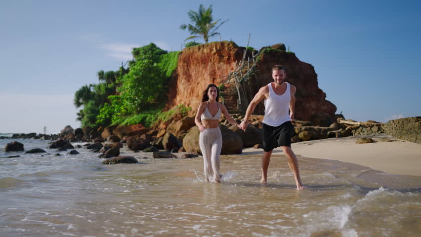 Young happy couple runs towards camera holding hands on beach through waves splashing water and enjoying summer. Boyfriend and girlfriend having fun at seaside in spectacular scenic tropical location. | Shutterstock HD Video #1101613941