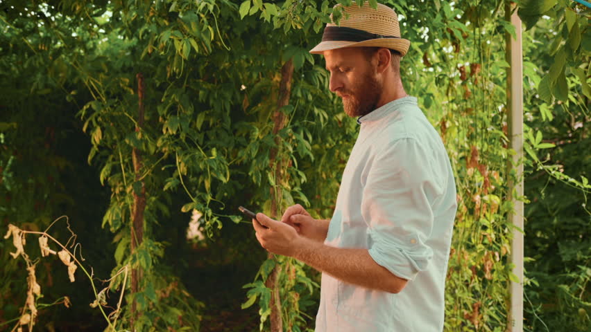 Contemporary agriculture. Bearded Caucasian man in a straw hat using smartphone. Slow motion. The concept of gardening and horticulture. | Shutterstock HD Video #1101615465
