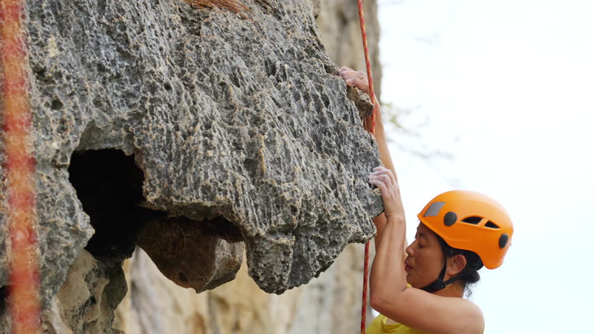 Asian mature woman climber with safety hanging rope climbing on rocky mountain at tropical beach island. Healthy people enjoy outdoor active lifestyle extreme sport climbing on summer holiday vacation