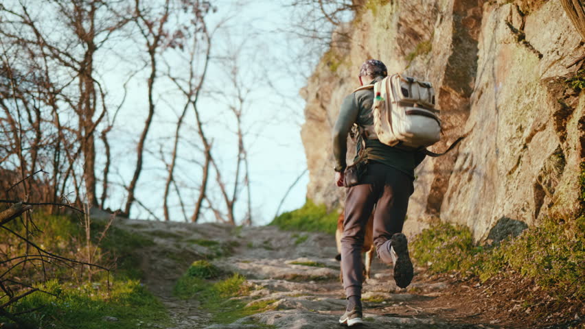 Slow motion shot of a man with a backpack running with his dog on a mountain path next to a rock. Back view