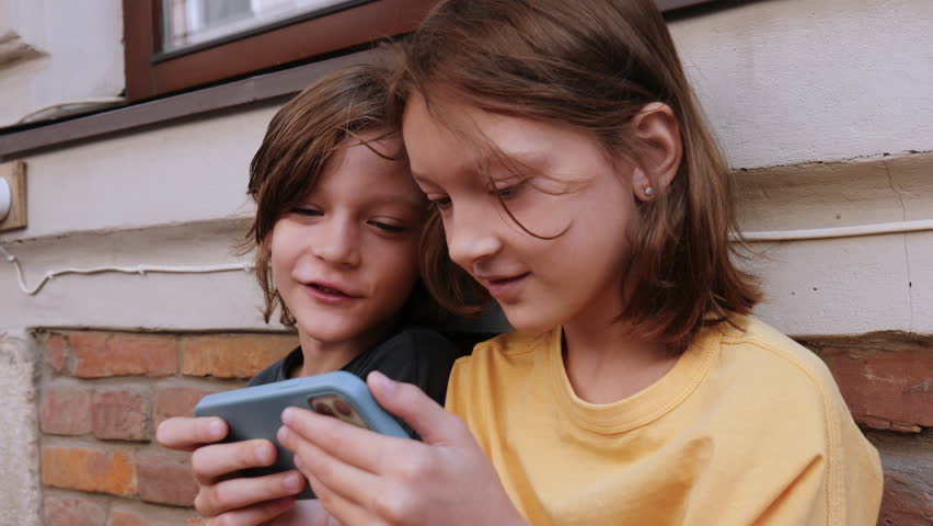 Two happy Caucasian teen age children, boy and girl playing fun mobile game together outside using phone in blue case.