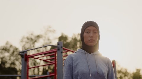 Portrait of a young asian muslim woman athlete in hijab and grey hoodie close up at the outdoor workout area with the setting sun in the background 스톡 비디오