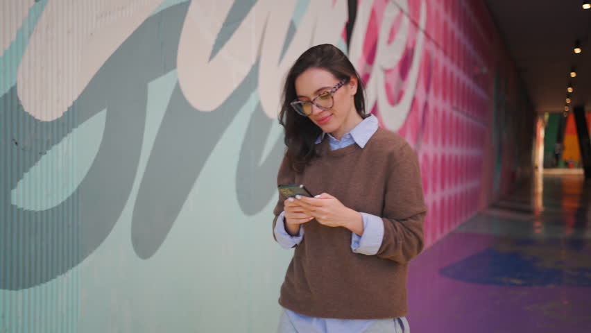 Woman using smartphone outdoor. Positive female person in glasses with leopard print use mobile phone outdoor. Happy attractive female freelancer or student with smartphone walking street Side view