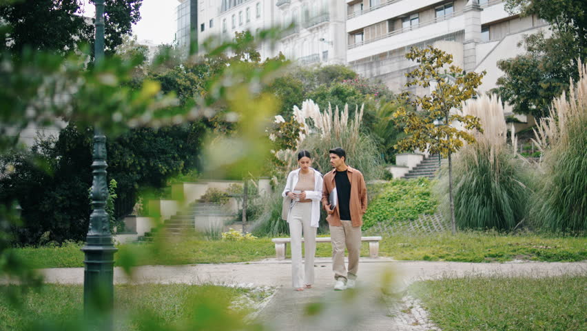 Confident colleagues crossing alley together. Successful businesscouple talking at green nature district. Serious woman holding take away coffee cup listening man. Two coworkers having work break 