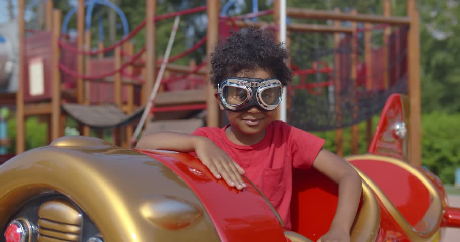 Portrait of adorable African-American toddler boy wearing pilot goggles sitting in toy plane on playground. Happy smiling African-American kid playing in summer park